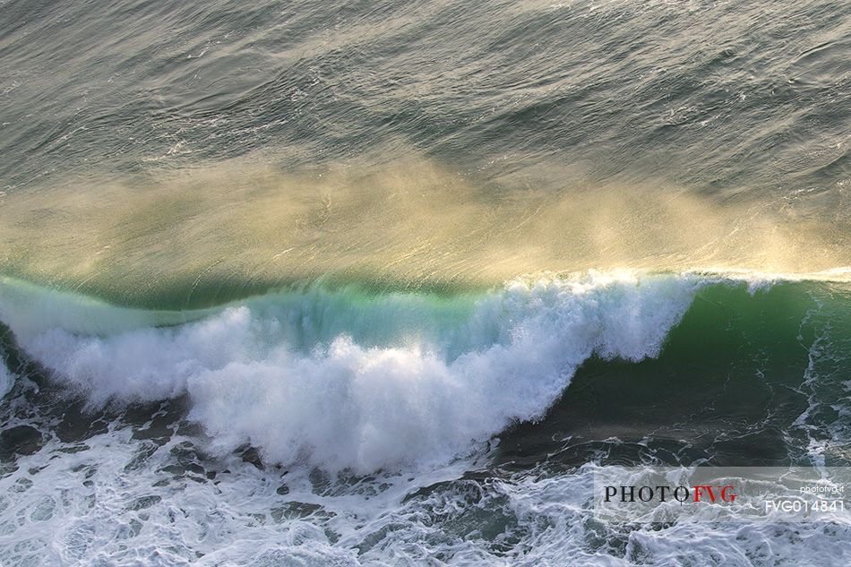 The sun at sunset, illuminates the waves crashing against the cliffs of Capo Nieddu