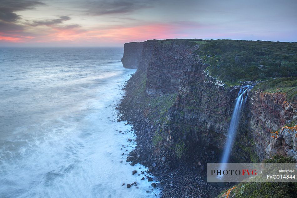 The sun at sunset, illuminates the waves crashing against the cliffs of Capo Nieddu where a magical waterfall plunges into the wonderful sea of Sardinia