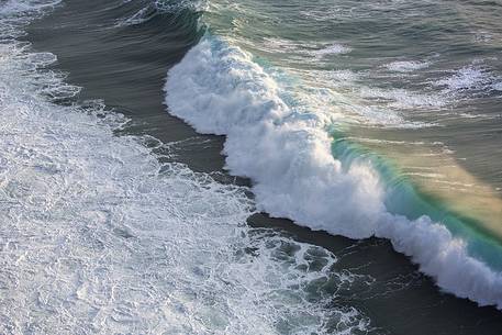 The sun at sunset, illuminates the waves crashing against the cliffs of Capo Nieddu