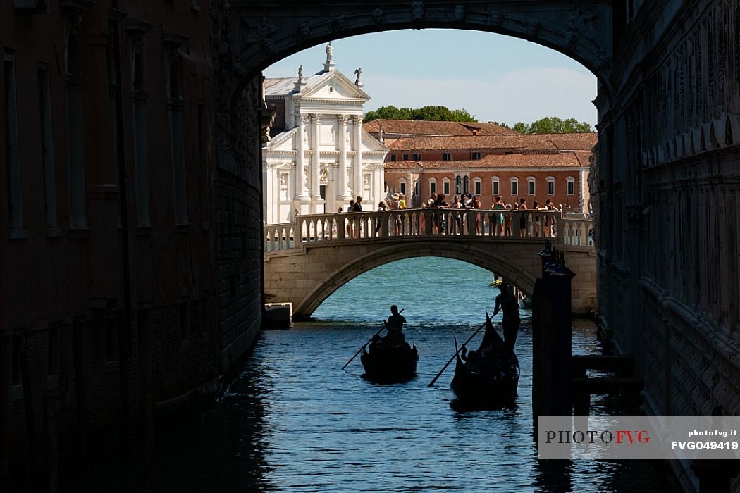 Gondoliers pass under the Bridge of Sighs, while tourists photograph them from the Ponte della Paglia. In the background, the facade of the church of San Giorgio Maggiore designed by Palladio. Venice