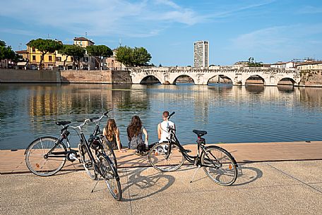 Roman Tiberius Bridge on the river Marecchia in Rimini and some people sit on the banks with their bikes, Rimini, Emilia Romagna, Italy, Europe