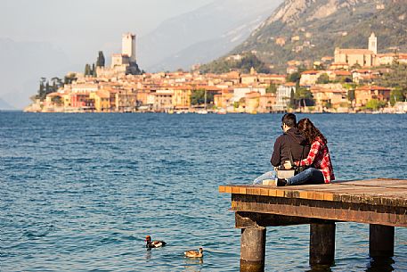 Couple sitting on a gangway in Garda lake in front of the small medieval village of Malcesine, Italy