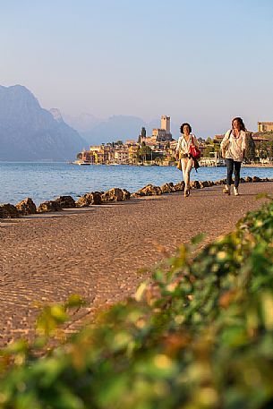 Two women walk along Garda lake at sunset, in the background the small medieval village of Malcesine, Italy