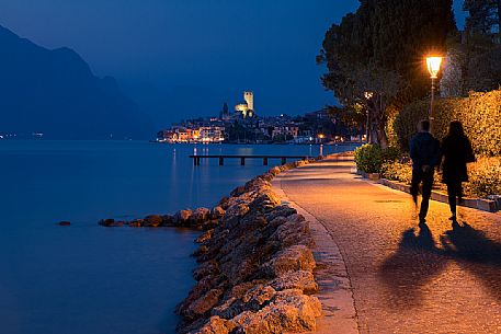 Evening walking along the shores of Garda lake, in the background the small medieval village of Malcesine with its illuminated Scaligero castle, Italy