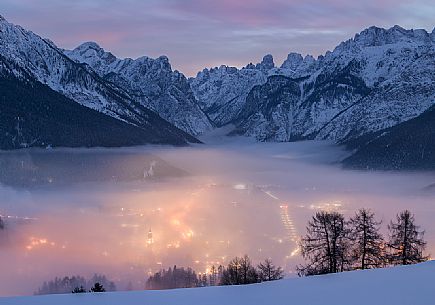 A dense fog dominates the village of Dobbiaco that lights up after sunset, in the background the peaks of Dobbiaco, Piz Popena and Monte Cristallo, Pusteria valley, dolomites, Trentino Alto Adige, Italy, Europe