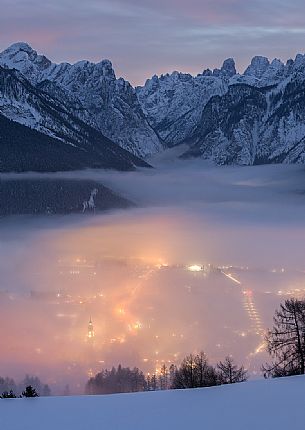 A dense fog dominates the village of Dobbiaco that lights up after sunset, in the background the peaks of Dobbiaco, Piz Popena and Monte Cristallo, Pusteria valley, dolomites, Trentino Alto Adige, Italy, Europe