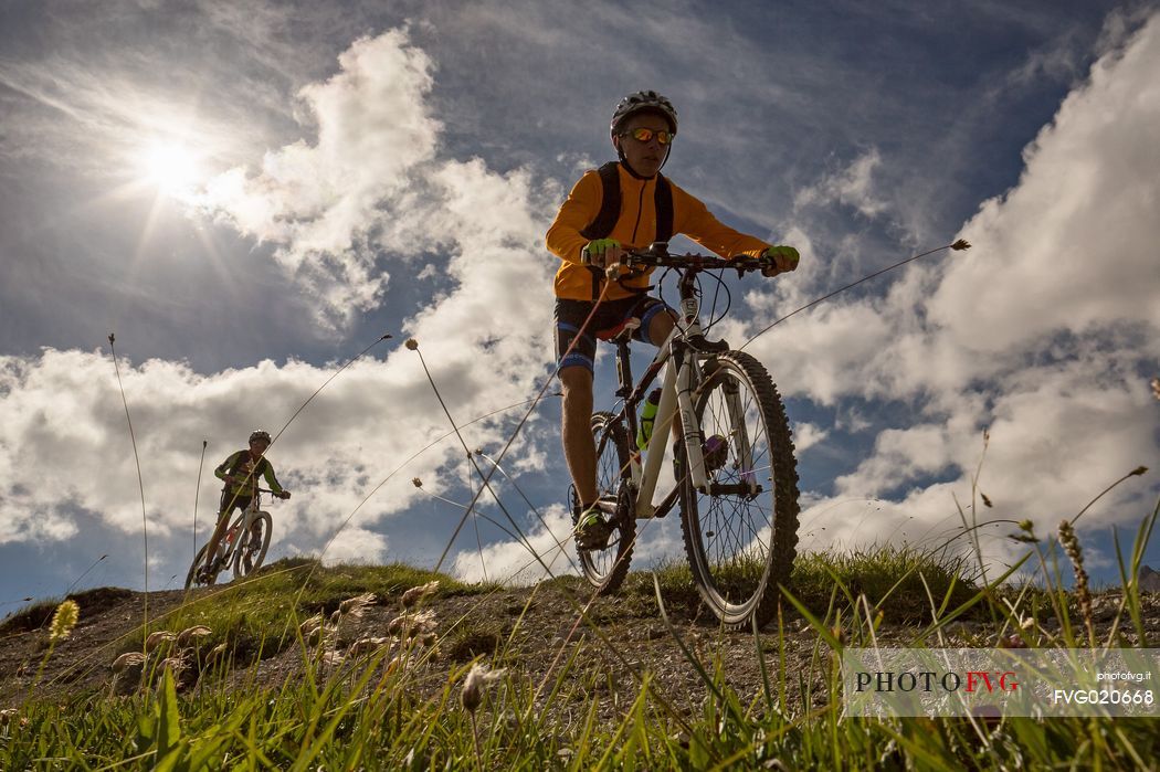 Two young mountain bikers come down from Biscia summit, in the background the Sesto Dolomites, Cadore, Italy