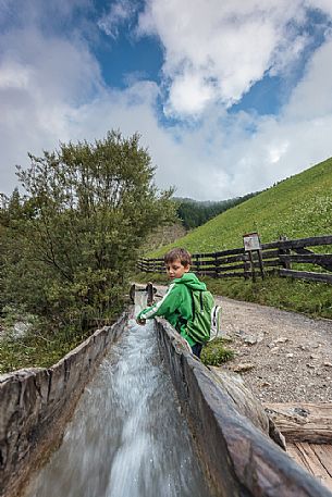 Child plays with natural water that flow in the traditional wooden canal near a water mill, Longiar, Badia valley, dolomites, Trentino Alto Adige, Italy, Europe