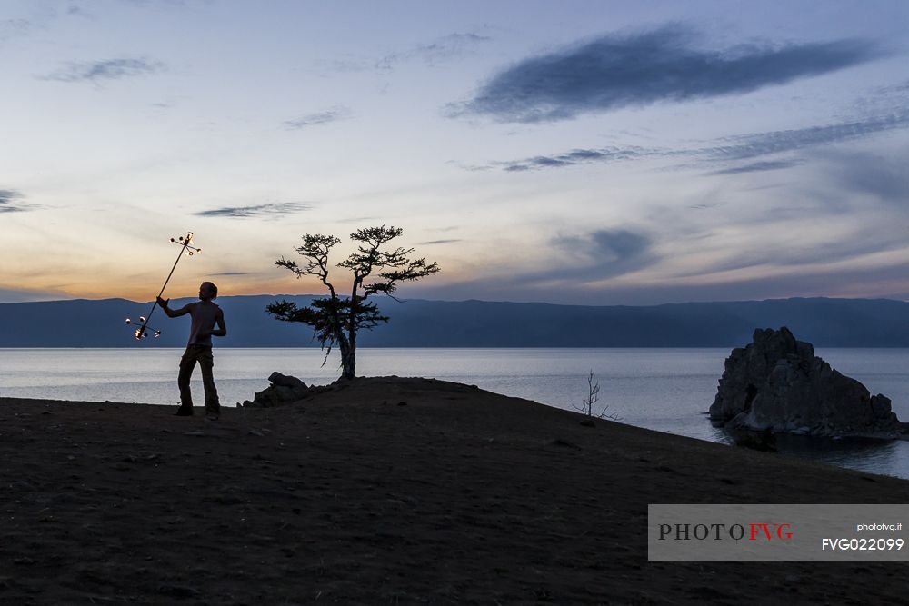 A boy dancing and playing with fire during the sunset at Olkhon Island. In the background the holy Shamanka or Shaman's Rock, Bajkal lake, Russia