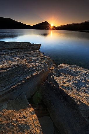 At dawn the sun peeks out behind the lake of Campotosto that seems to be preparing for a day full of light, Gran Sasso and monti della Laga national park, Abruzzo, Italy At dawn the sun peeks out behind the lake of Campotosto that seems to be preparing for a day full of light, Gran Sasso and monti della Laga national park, Abruzzo, Italy