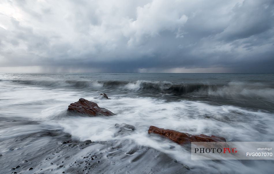 The cold water of the English Channel vigorously hits a rocky beach on the Normandy coast, while in the distance a powerful storm unleashes pouring rain and dark clouds upon the sea.