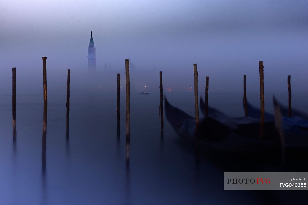 On a foggy winter morning, the bell tower of the San Giorgio island breaks through the fog while the gondolas sway slightly in the Venice lagoon, Venice, Veneto, Italy