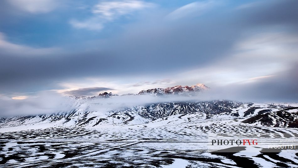 Clouds blown by the wind flow veiling the mountains lit by the last light of a winter sunset, Campo Imperatore, Gran Sasso e Monti della Laga National Park