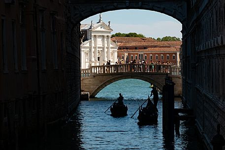 Gondoliers pass under the Bridge of Sighs, while tourists photograph them from the Ponte della Paglia. In the background, the facade of the church of San Giorgio Maggiore designed by Palladio. Venice