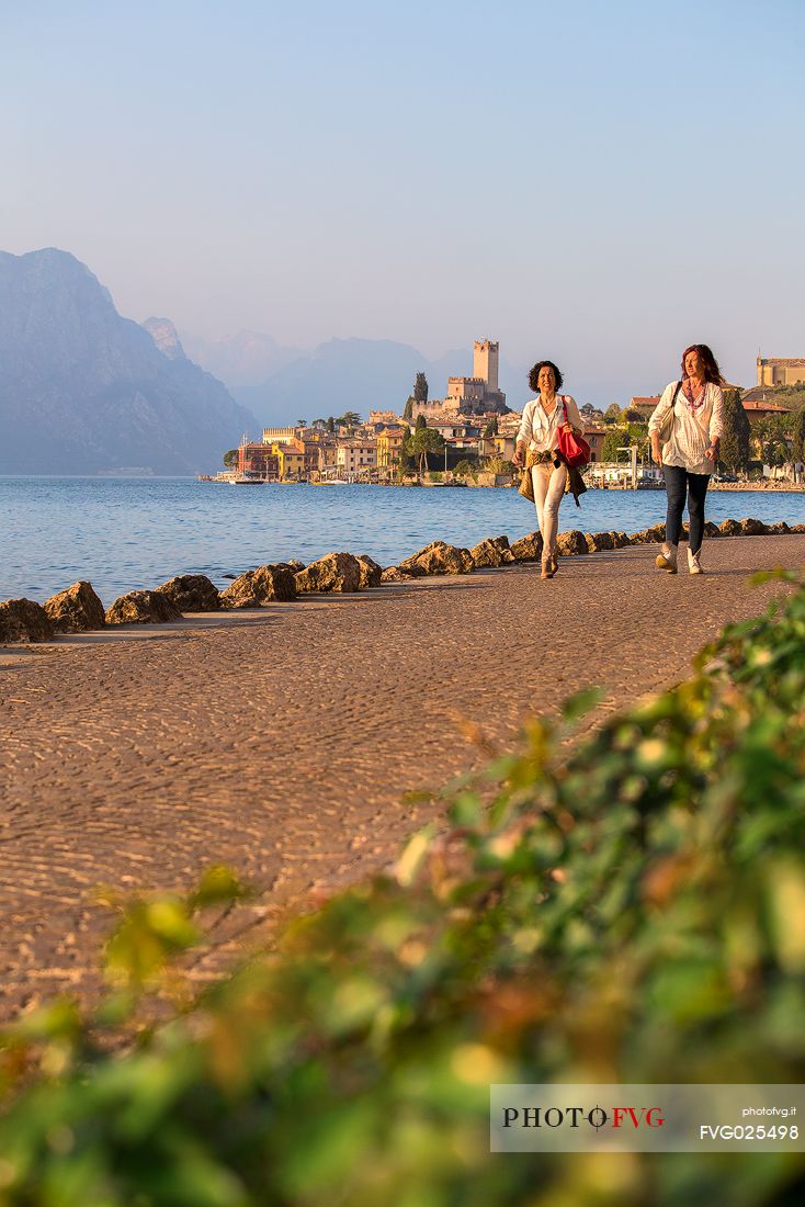 Two women walk along Garda lake at sunset, in the background the small medieval village of Malcesine, Italy