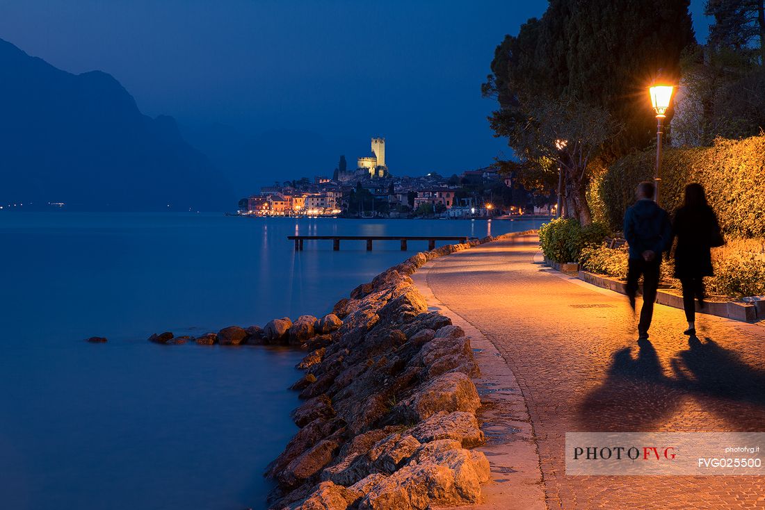 Evening walking along the shores of Garda lake, in the background the small medieval village of Malcesine with its illuminated Scaligero castle, Italy
