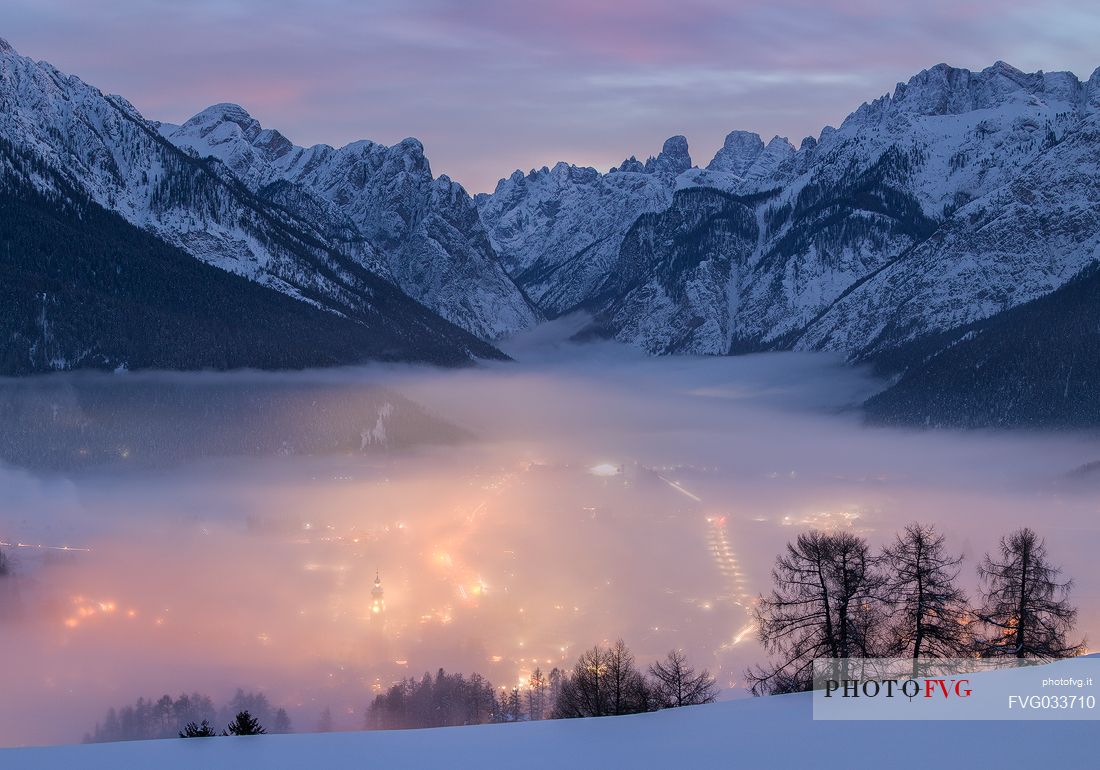 A dense fog dominates the village of Dobbiaco that lights up after sunset, in the background the peaks of Dobbiaco, Piz Popena and Monte Cristallo, Pusteria valley, dolomites, Trentino Alto Adige, Italy, Europe