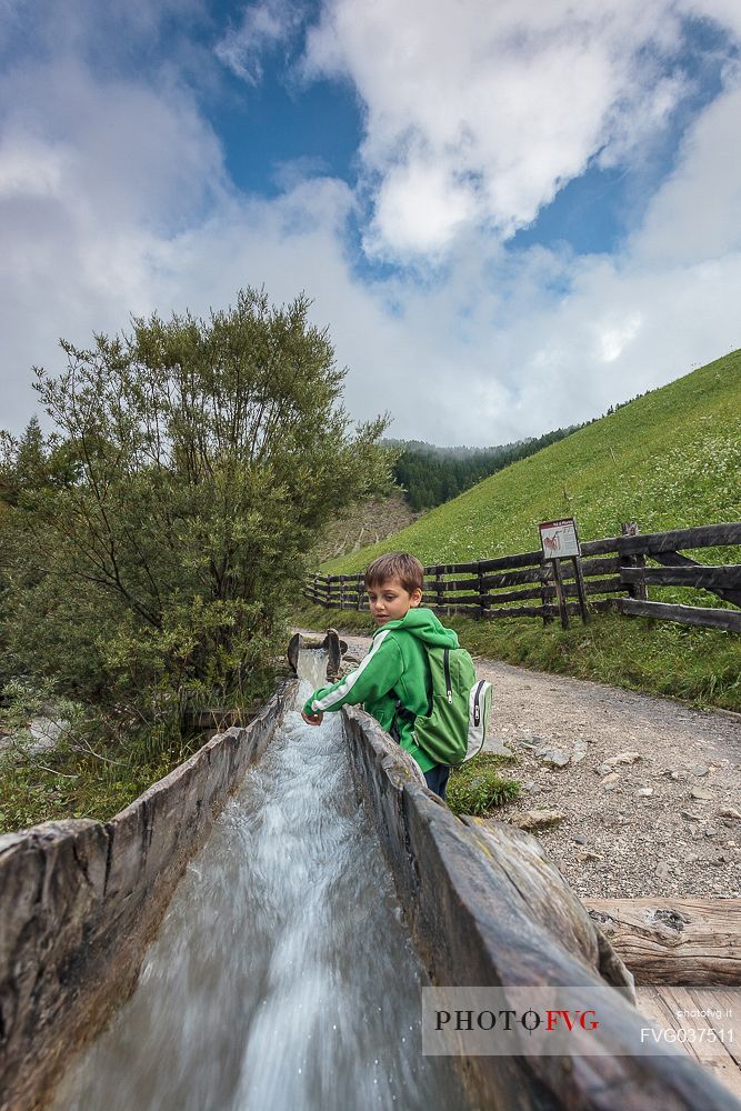 Child plays with natural water that flow in the traditional wooden canal near a water mill, Longiar, Badia valley, dolomites, Trentino Alto Adige, Italy, Europe