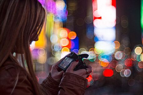 Young woman watches photo taken in her camera, in the background the night lights of Times Square, Manhattan, New York, United States