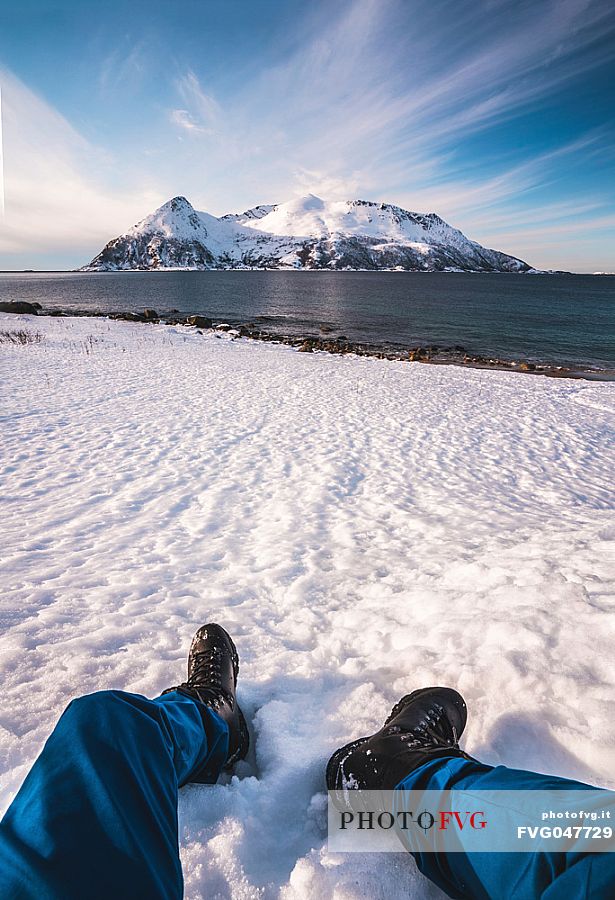 A hiker rests on the beach of Rekvik,a small village near Tromvik, in the background the Sessoya island, Tromso, Norway, Europe
