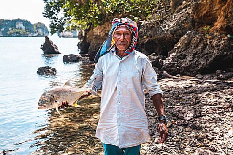 Portrait of senior fisherman holding a fish in a beautiful bay of Wigeo Island, Raja Ampat archipelago, West Papua, Indonesia