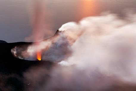 An explosion produces a fountain of lava and lapilli. In the background the sunset is reflected on the sea